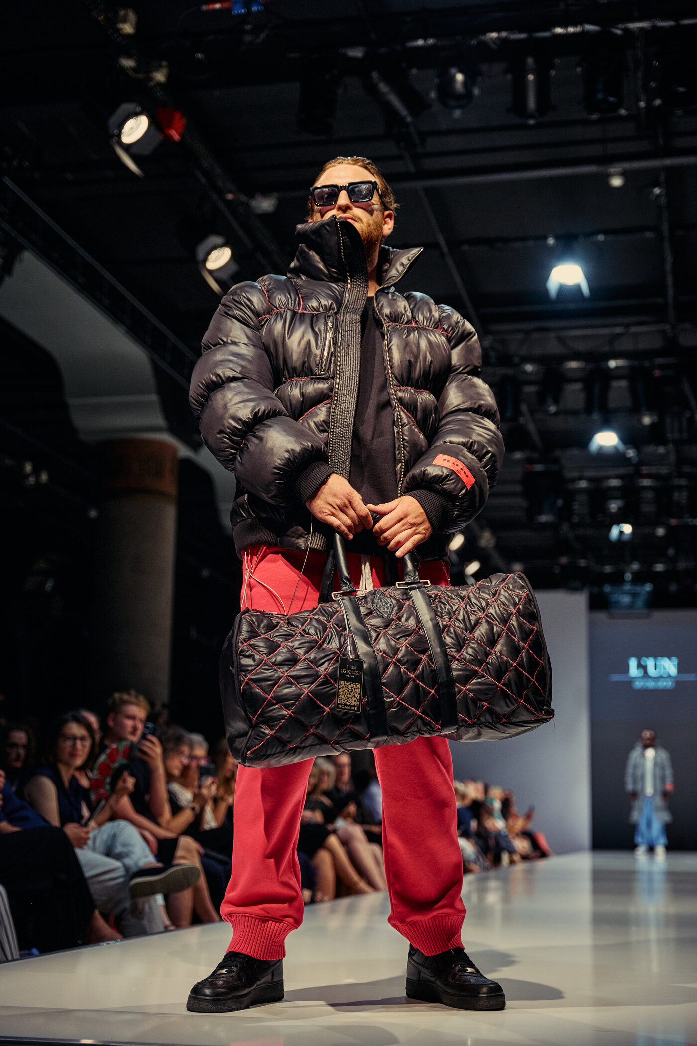 Look 01 „Éclat Noir“ — schwarze Glanz-Pufferjacke mit roten Kontrastnähten, rote Jogginghose und gesteppter Weekender auf der Mannheimer Runway
